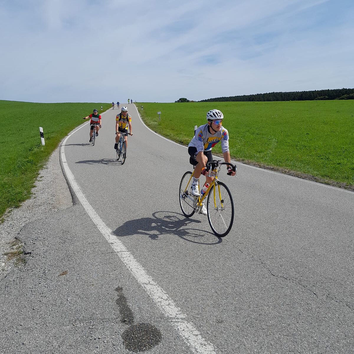 Drei Rennradfahrende auf Landstraße grüne Wiese links und rechts der Straße
