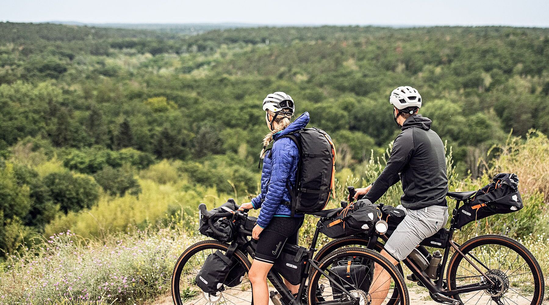 Zwei Radfahrende mit Gravelbikes und Gepäck stehen auf einer Anhöhe und blicken auf einen weiten Wald. 