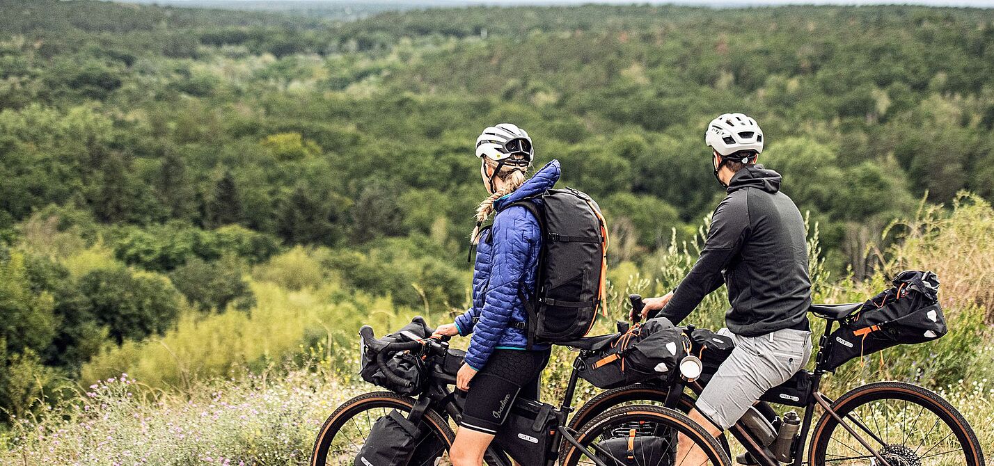 Bikepacking Artikel Zwei Radfahrende mit Gravelbikes und Gepäck stehen auf einer Anhöhe und blicken auf einen weiten Wald.